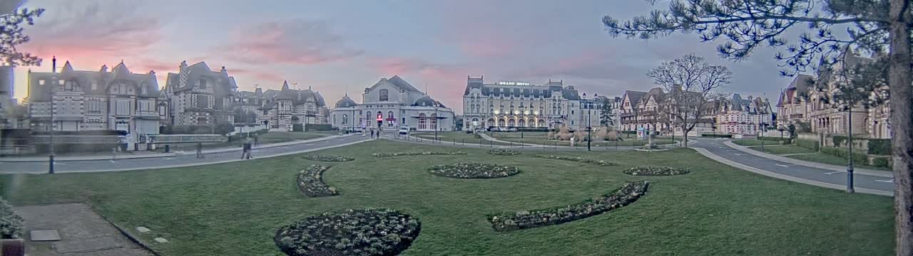 A grassy town square with ornate buildings and a large hotel is seen at dusk under a pale pink sky.