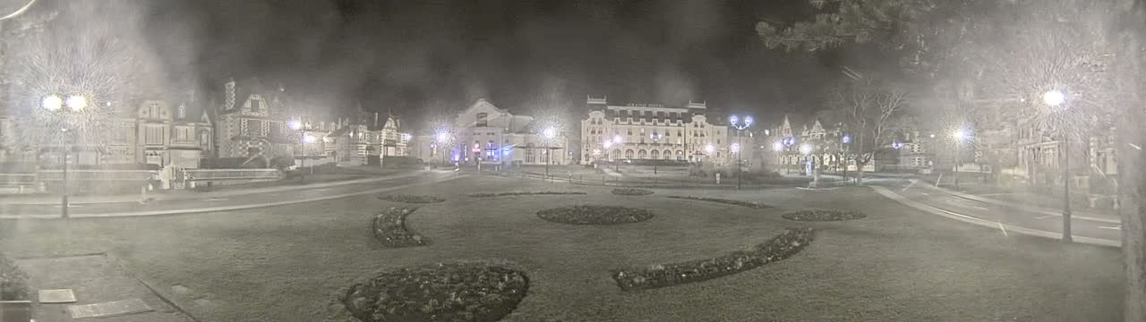 A nighttime view of a town square with several buildings, illuminated by streetlights, under snowy conditions.