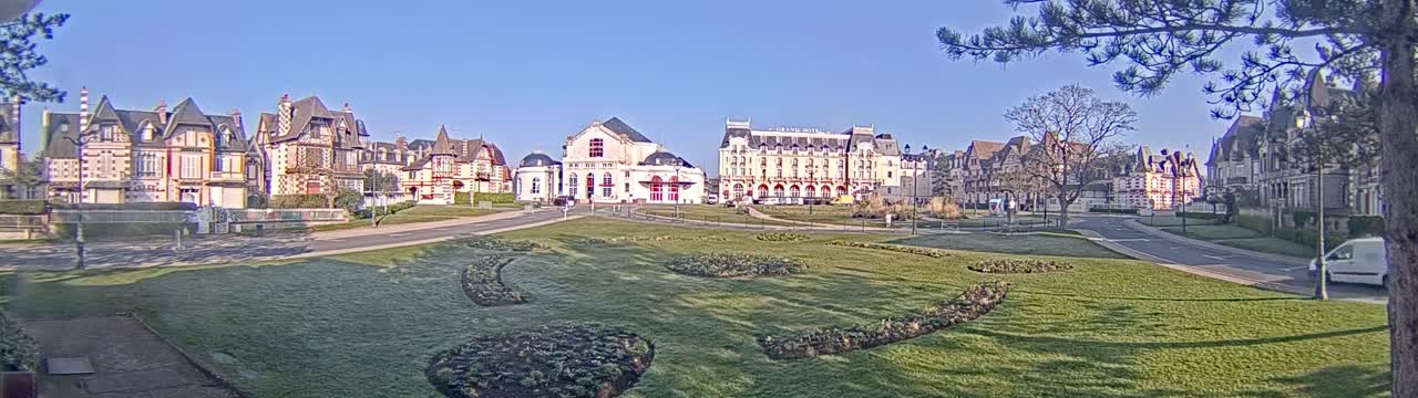 A grassy park with geometrically-shaped flowerbeds sits in front of a row of ornate buildings on a sunny day.