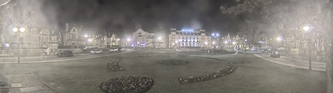 A nighttime view of a town square with several buildings, cars, and landscaped areas, under hazy or foggy conditions.