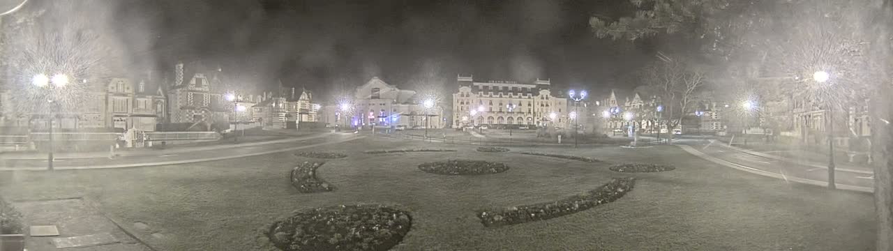 A snow-covered town square at night, illuminated by streetlights, features several buildings and landscaped flowerbeds.