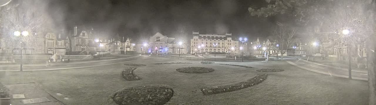 A snow-covered town square at night, illuminated by streetlights, features several buildings, including a large hotel, and landscaped areas with curved flowerbeds.