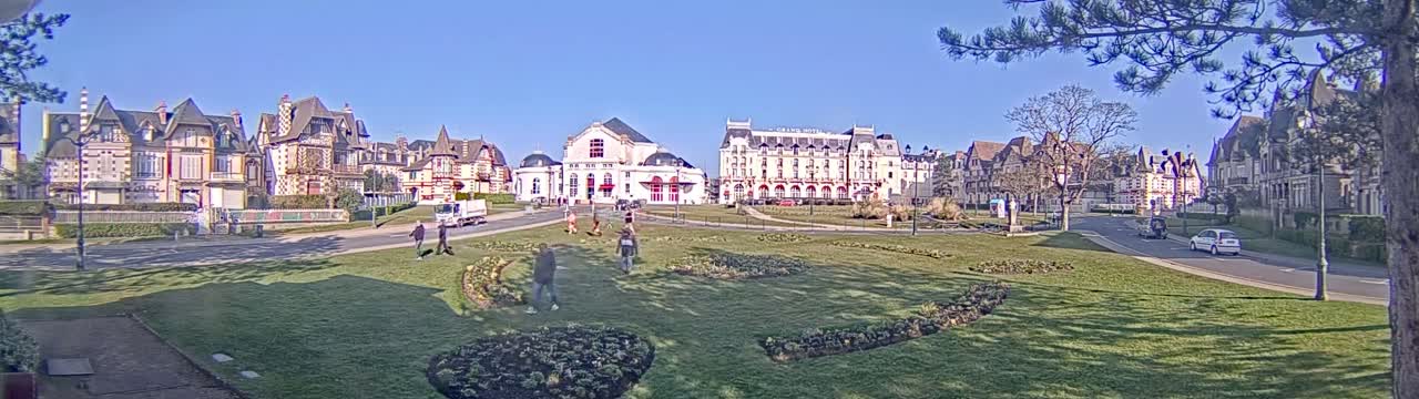 A sunny day reveals a grassy park with several pedestrians, surrounded by a row of ornate buildings, including a large hotel.