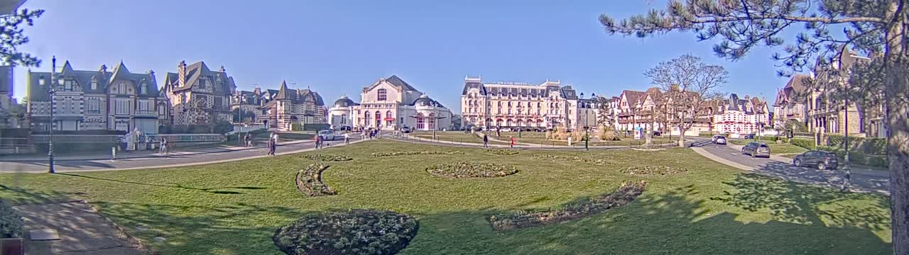 A grassy town square with several large buildings and a few people walking around on a sunny day.