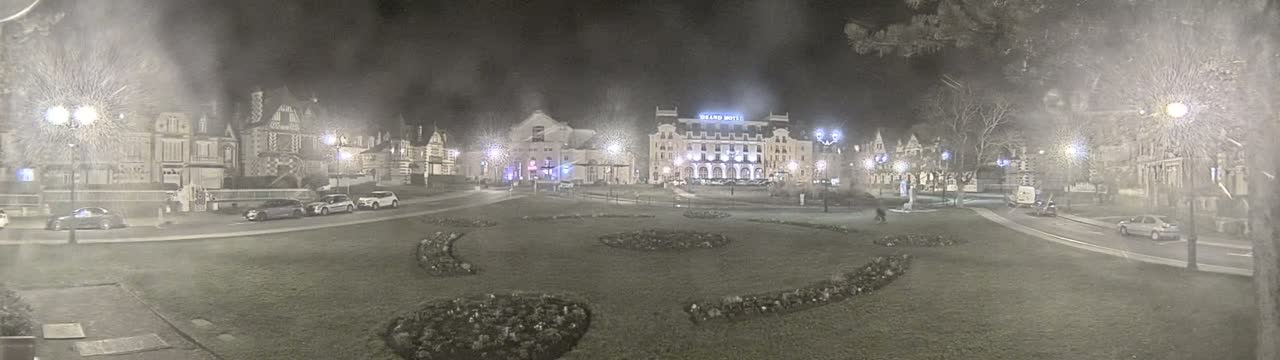 A nighttime view of a town square with several buildings, cars parked along the sides, and landscaped areas with flowerbeds.