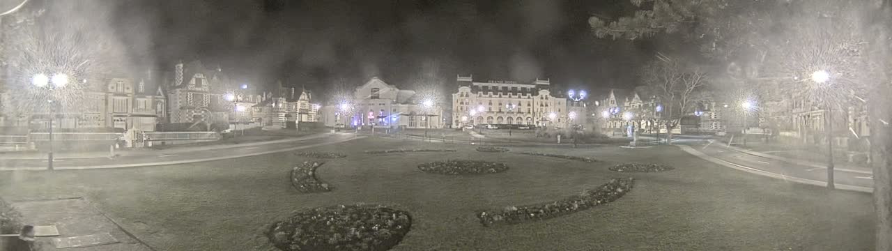 A nighttime view of a town square with several buildings, including a large hotel, surrounded by a grassy area with landscaped flowerbeds, under a hazy or snowy sky.