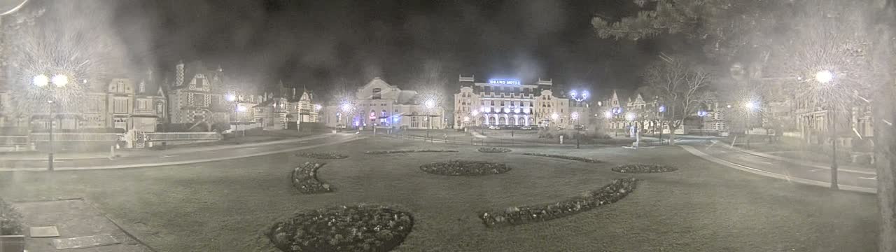A nighttime view of a town square with several buildings, including a large hotel, surrounded by landscaped gardens under hazy or snowy conditions.