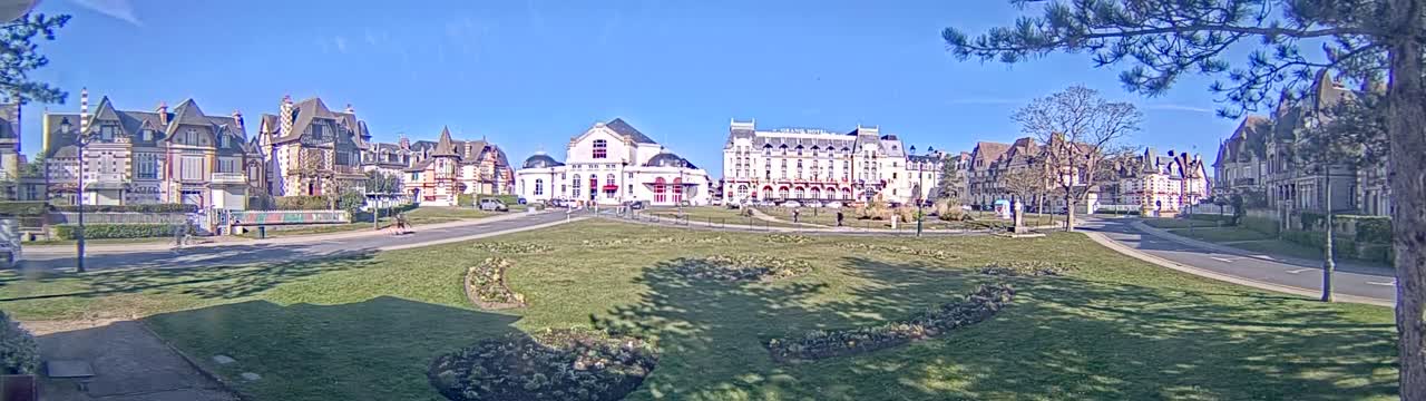 A sunny day reveals a row of ornate buildings and a large hotel across a grassy square with curving roads.