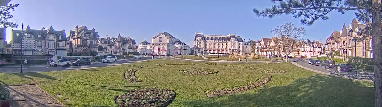 A sunny day reveals a grassy plaza with flowerbeds, surrounded by several ornate buildings and parked cars.