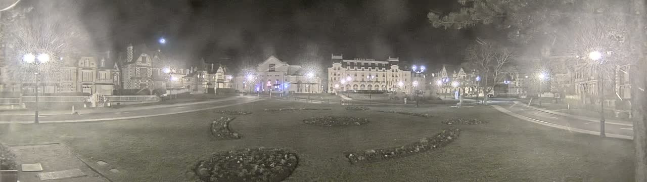 A nighttime view of a town square with several buildings, lit by street lamps, under hazy conditions.