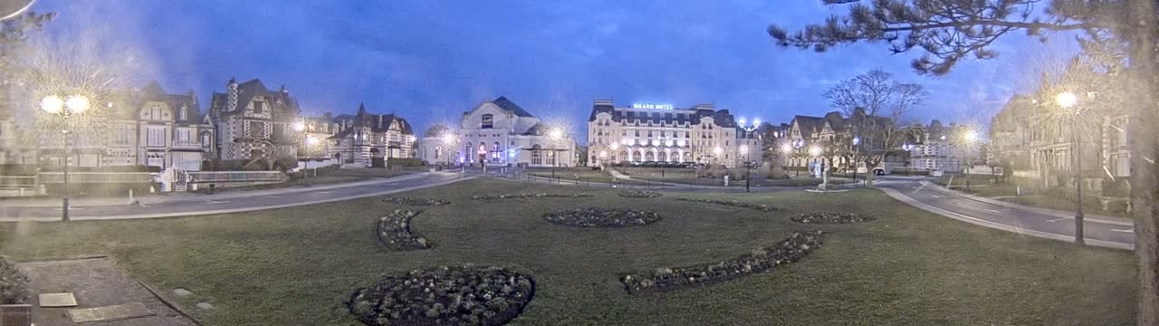 A town square at twilight, with several large buildings, grassy areas containing decorative landscaping, and streetlights illuminating the scene.