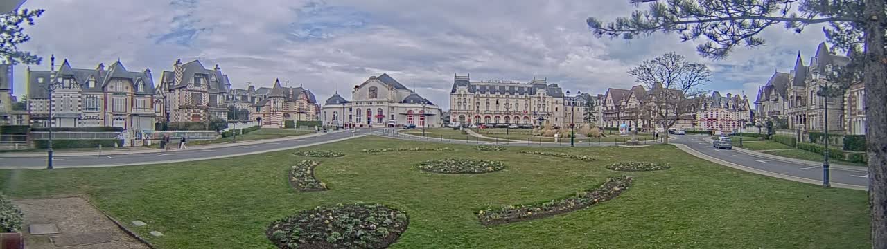 A grassy plaza with flowerbeds is centered in front of a row of ornate buildings under a cloudy sky.