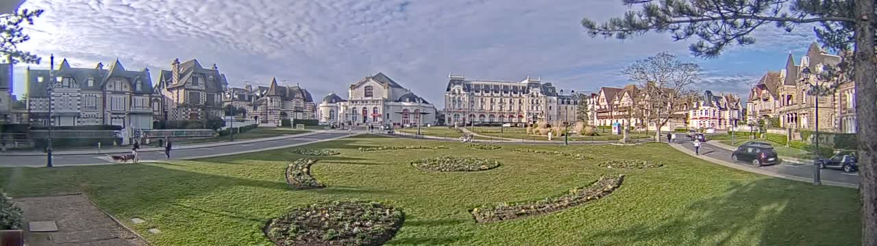 A grassy park with geometric flowerbeds is situated in front of several large, ornate buildings under a partly cloudy sky.