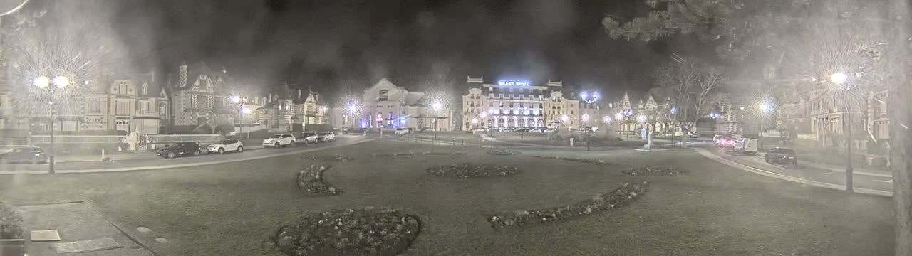 A nighttime view of a town square with several buildings, including a large hotel, and parked cars, under overcast conditions.