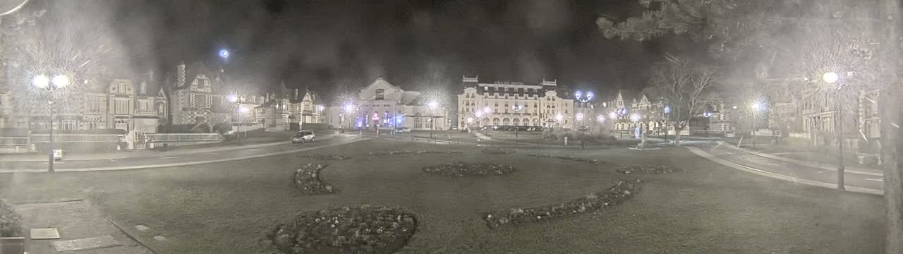 A nighttime view of a town square with several buildings, including a large hotel, surrounded by a grassy area with landscaping, under overcast conditions.