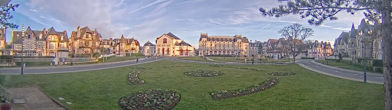 A grassy park with flowerbeds is in front of a row of large, ornate buildings under a partly cloudy sky.