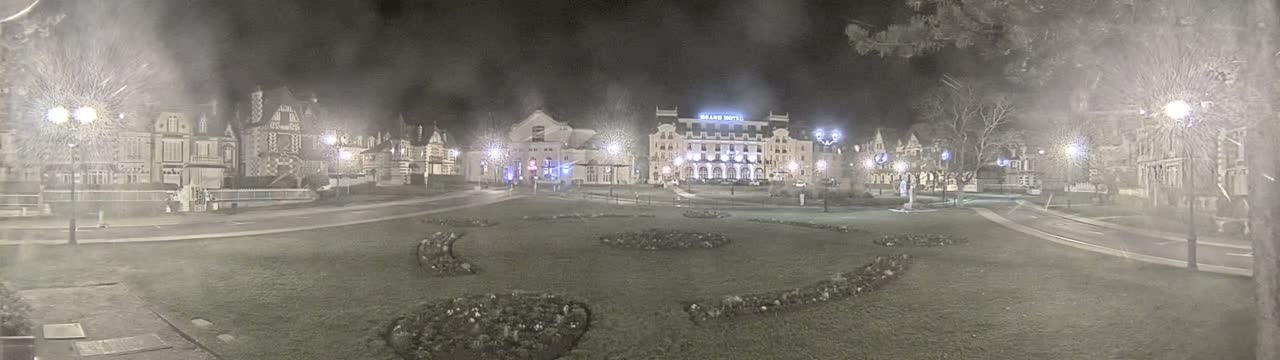 A town square at night, under a hazy sky, is surrounded by buildings and features several flowerbeds.