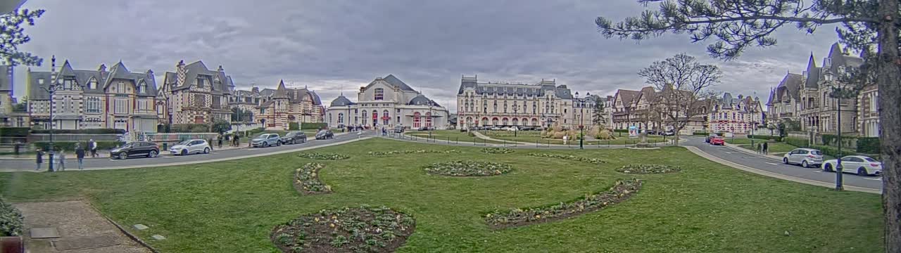 A grassy square with flowerbeds is situated in front of several ornate buildings, under a cloudy sky.