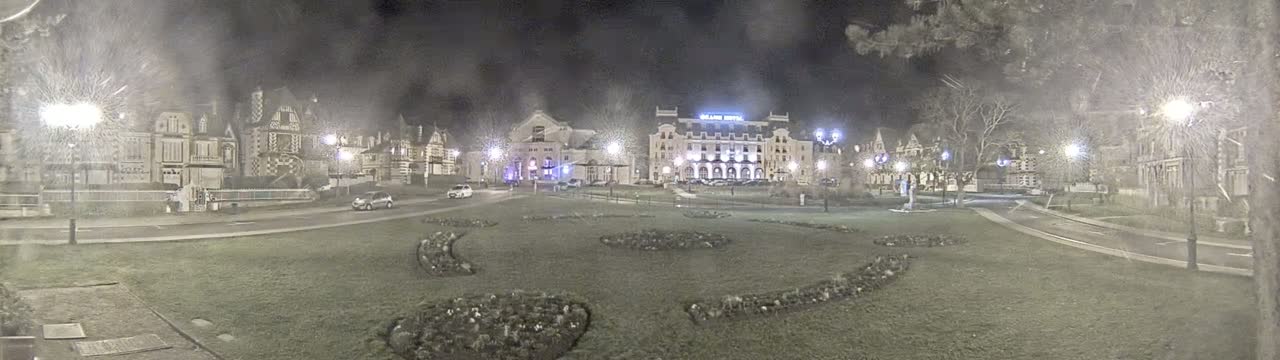 A nighttime view of a town square with several buildings, including a large hotel,  lit by streetlights under overcast conditions.