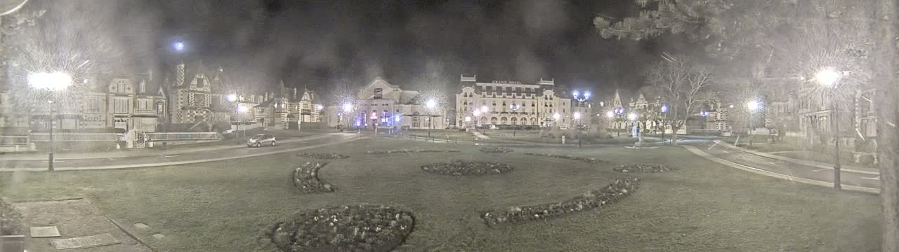 A nighttime scene of a town square with several buildings, a car, and landscaping, lightly snowing.