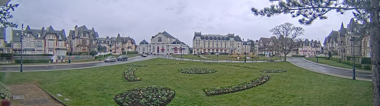 A grassy town square with flowerbeds is surrounded by ornate buildings under an overcast sky.