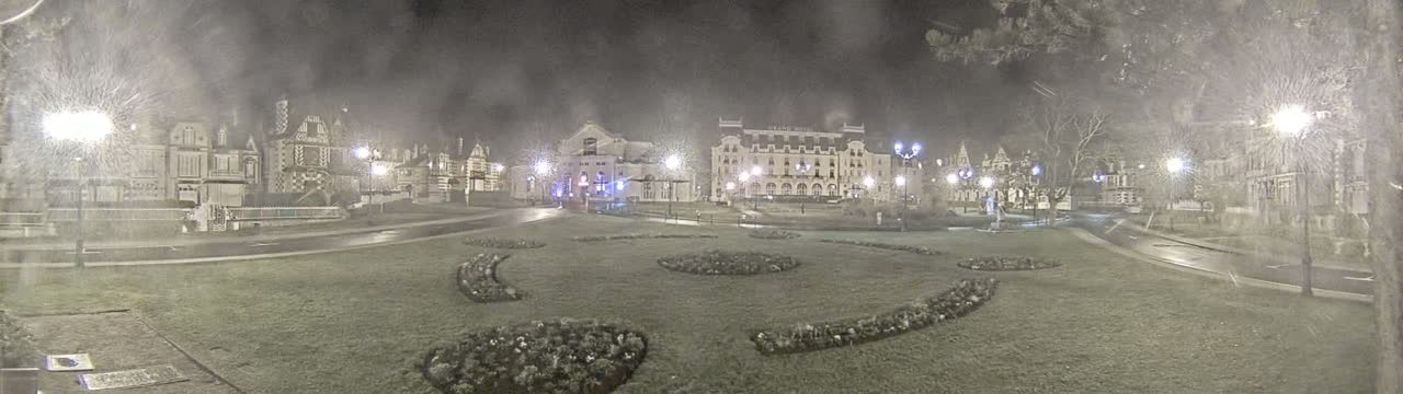 A nighttime view of a town square with several buildings, including a large hotel, surrounded by snow-covered grass and landscaping.