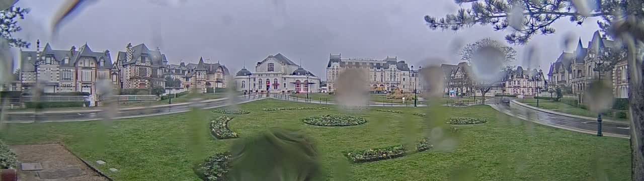 A rain-slicked town square with ornate buildings and meticulously landscaped flowerbeds sits under an overcast sky.