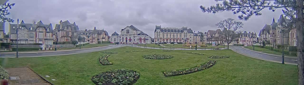 A grassy plaza with ornate flowerbeds is surrounded by several large, ornate buildings under an overcast sky.
