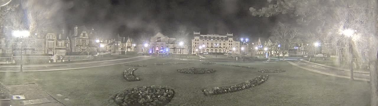 A nighttime view of a town square with several buildings, including a large hotel, surrounded by landscaped areas and lit by streetlights in snowy conditions.