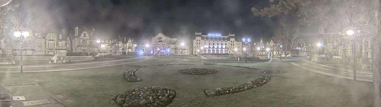 A nighttime view of a town square with several buildings, including a large hotel, surrounded by landscaped areas under hazy conditions.