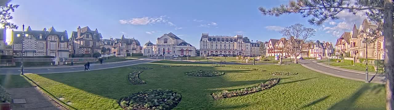 A sunny day reveals a row of elegant, multi-story buildings with ornate facades surrounding a grassy park featuring landscaped flower beds and curving pathways.