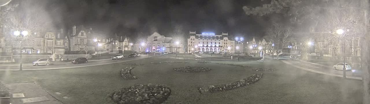 A nighttime view of a town square with several buildings, cars, and a grassy area with flowerbeds, under slightly hazy conditions.