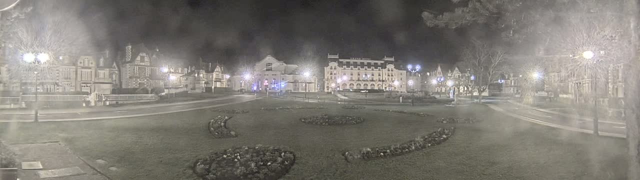 A nighttime scene shows a town square with several buildings, including a large hotel, surrounded by a grassy area with landscaped flowerbeds, under hazy, possibly snowy or foggy, conditions.