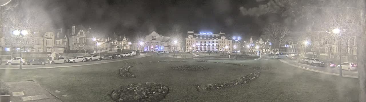 A nighttime view of a town square with several buildings, including a large hotel, surrounded by parked cars and landscaped areas under hazy conditions.
