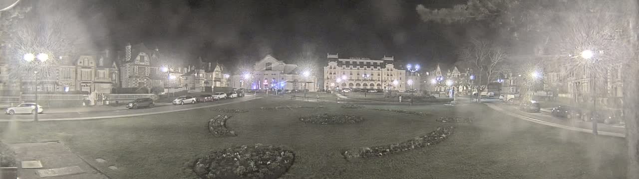 A nighttime, hazy view of a town square with several buildings, including a large hotel, cars parked along the street, and landscaping featuring curved flower beds.