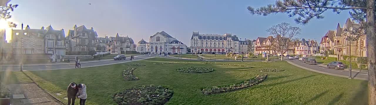 A sunny day reveals a grassy park with flowerbeds, surrounded by several large, ornate buildings and cars parked along nearby streets.
