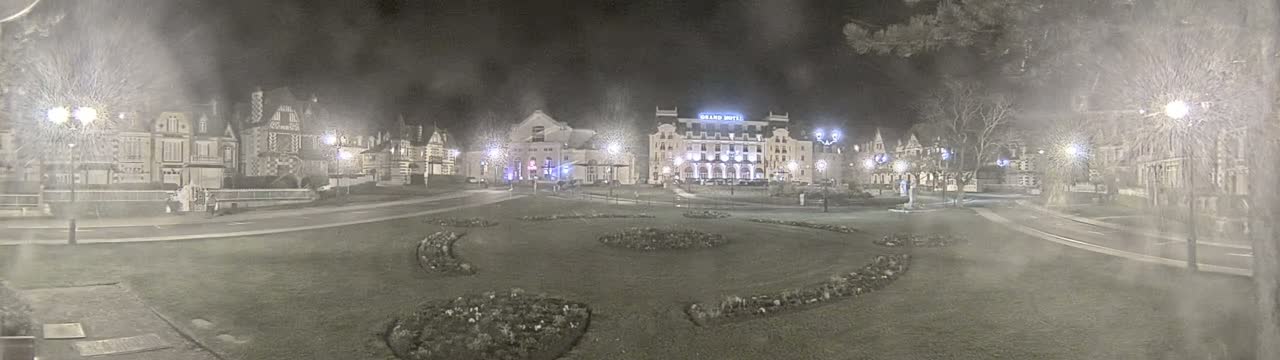 A snow-covered town square at night, illuminated by streetlights, features several buildings, including a large hotel, and landscaped flowerbeds.