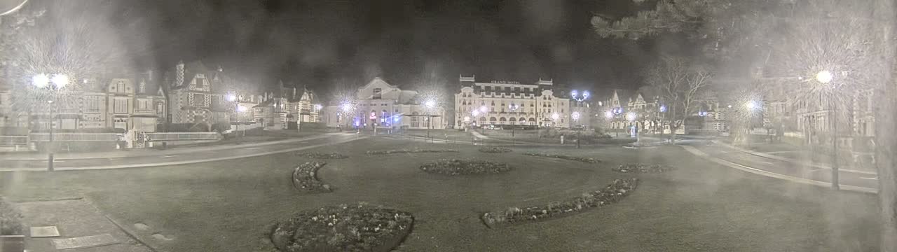 A nighttime view of a town square with several buildings, including a large hotel, surrounded by snow-covered lawns and trees under hazy, snowy conditions.