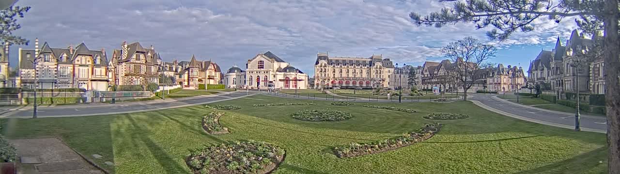 A wide, grassy park area with manicured flower beds is situated in front of several large, ornate buildings under a partly cloudy sky.