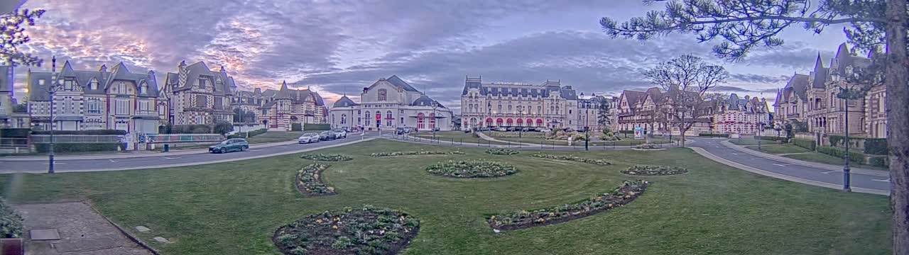 A town square with ornate buildings and flowerbeds is under a partly cloudy, twilight sky.