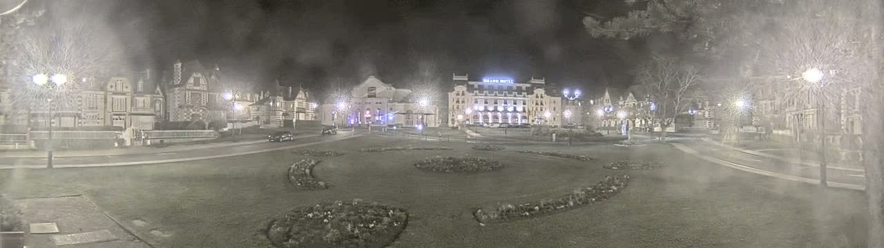 A nighttime view of a town square with several buildings, including a large hotel, surrounded by a grassy area with flowerbeds, under hazy or snowy conditions.