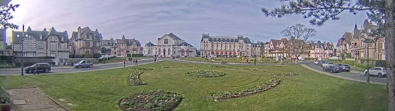 A town square with several ornate buildings, a grassy area with flowerbeds, and parked cars under a cloudy sky.