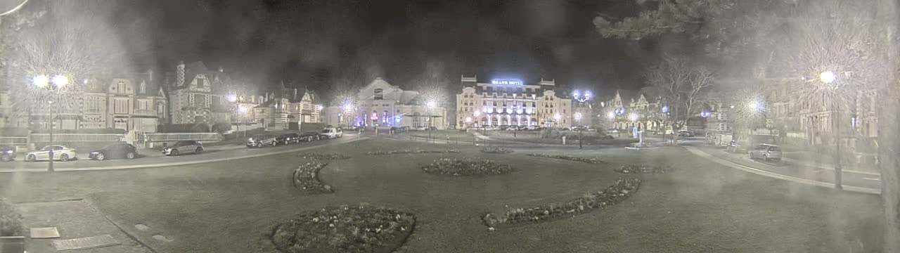 A nighttime view of a town square with several buildings, cars parked along the sides, and landscaped areas with low-lying plants, under snowy conditions.