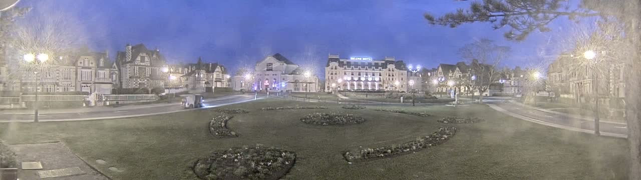 A town square at twilight, with several large buildings lining the perimeter, is surrounded by a grassy area with landscaped flower beds.