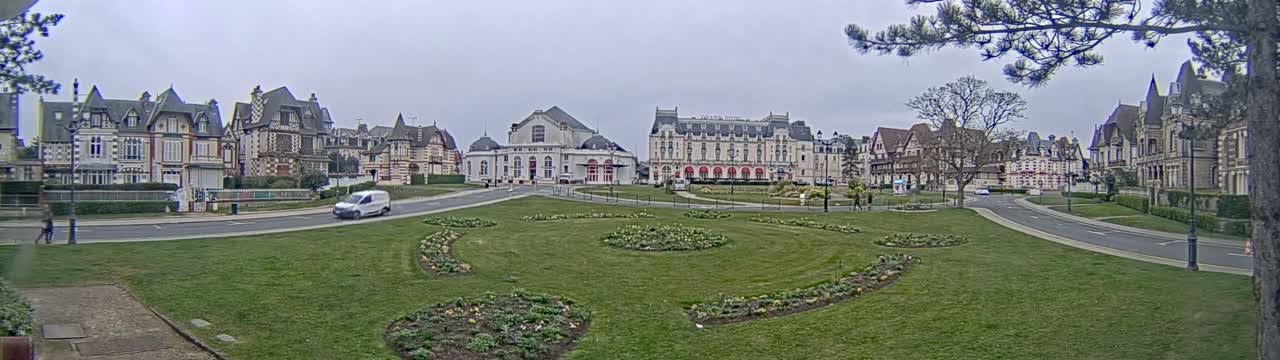 A grassy town square with flowerbeds is surrounded by several large, ornate buildings under an overcast sky.
