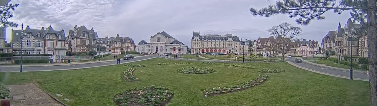 A grassy plaza with flowerbeds sits in front of a row of ornate, multi-story buildings under a cloudy sky.