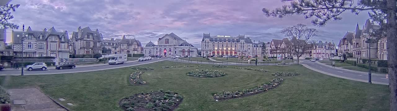 A row of ornate buildings and a large hotel are situated across a grassy square under a twilight sky.