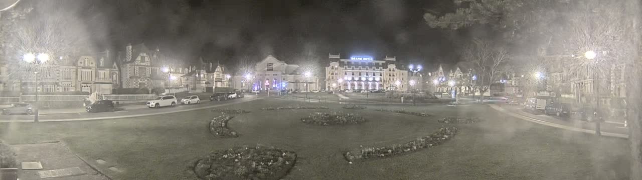 A nighttime view of a town square with several buildings, including a large hotel, surrounded by cars parked along the street, and flowerbeds in the center of the square, under slightly hazy conditions.