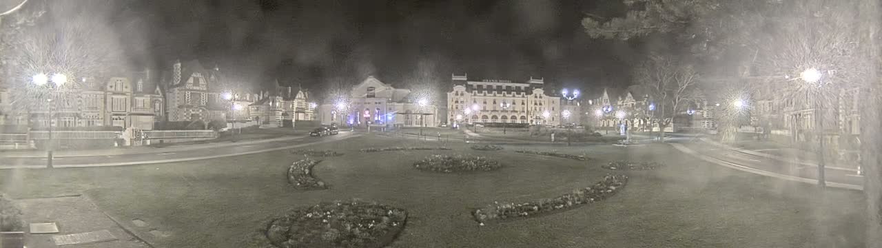 A nighttime view of a town square with several buildings, including a large hotel, illuminated by streetlights under a hazy sky.