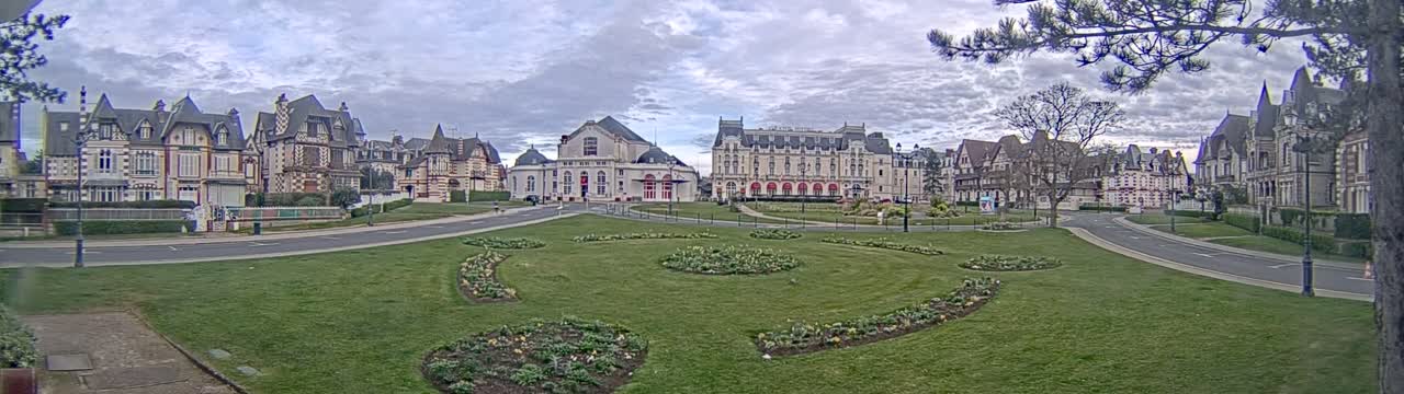 A grassy park with flowerbeds is in the foreground of a row of ornate buildings under a cloudy sky.